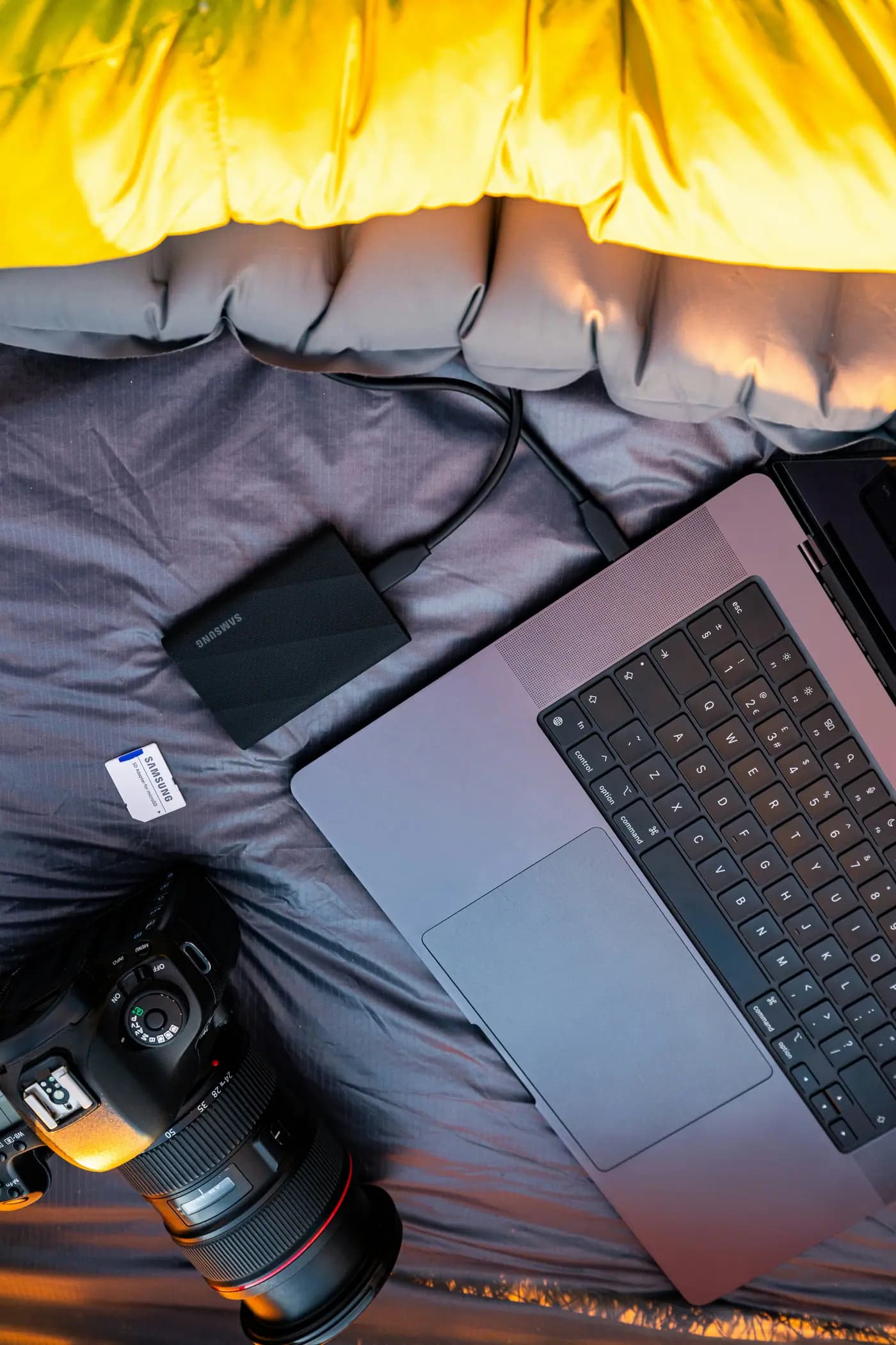 Laptop, camera, and SSD on a sleeping bag under a yellow blanket.