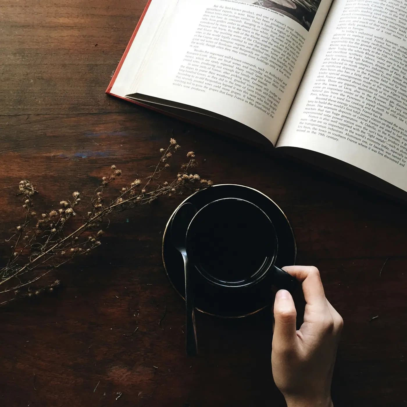 Hand holding a cup of coffee beside an open book on a wooden desk.